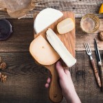 Close up hand holding cutting board with cheese, snack food on rustic table viewed from above