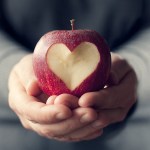 Hands holding an apple with heart shape carved into it.