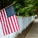 Small American flag hangs from a picket fence along the sidewalk in a rural town.