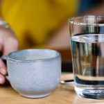 Hand holding a cup of coffee served with a glass of water and a spoon on a wooden plate on a wooden table