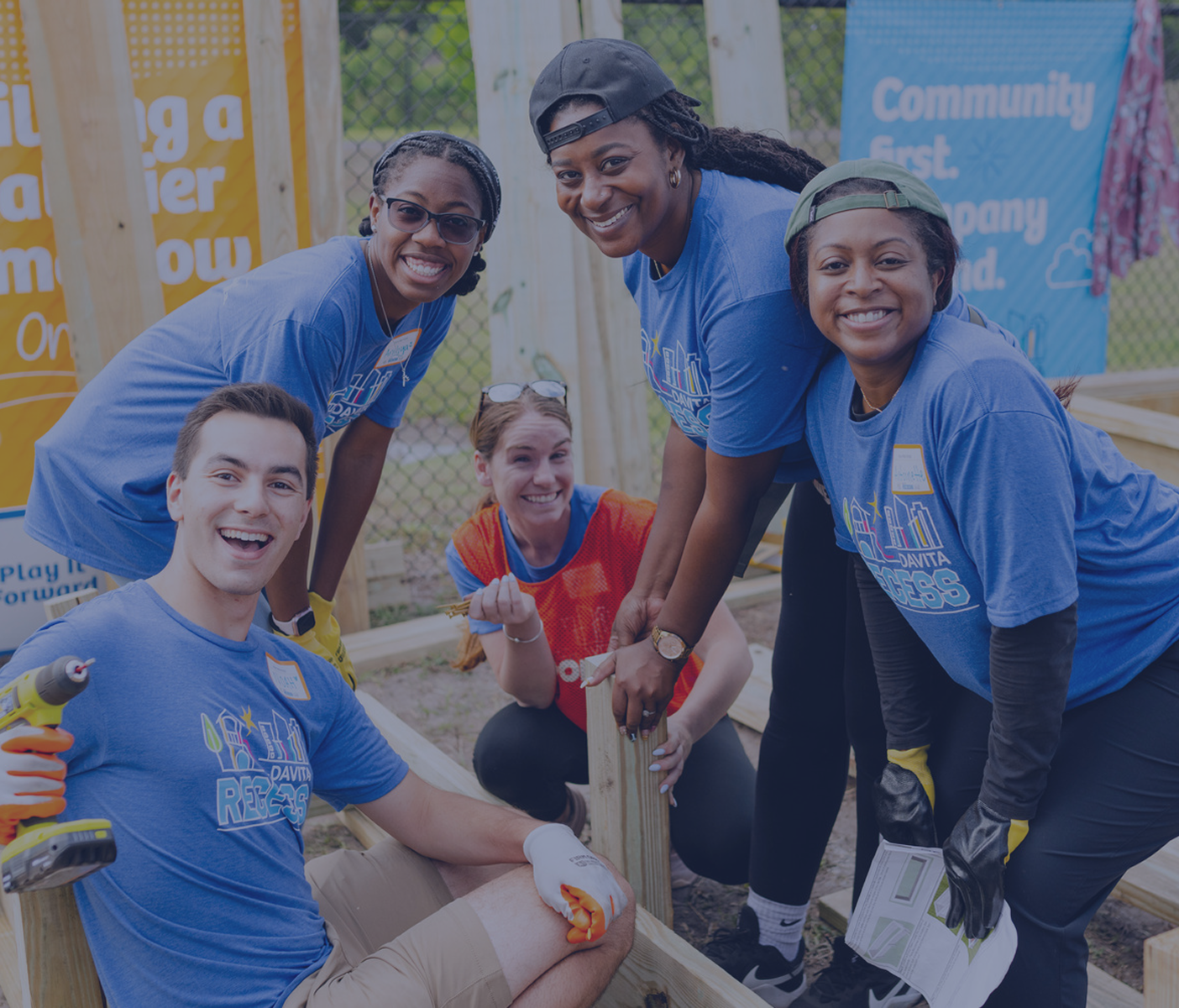 Five smiling volunteers in blue DaVita t-shirts working together with tools to build an outdoor wooden structure.