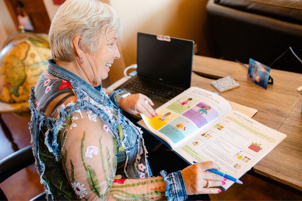 A smiling older woman sitting at a desk with a laptop, looking happily at an open book while holding a blue pen.