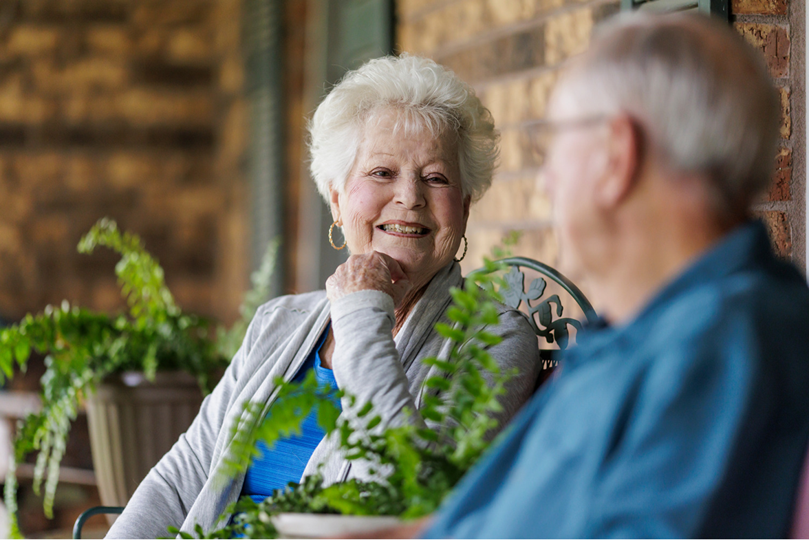 A smiling elderly woman sitting on a porch, looking happily at an older man seated next to her.