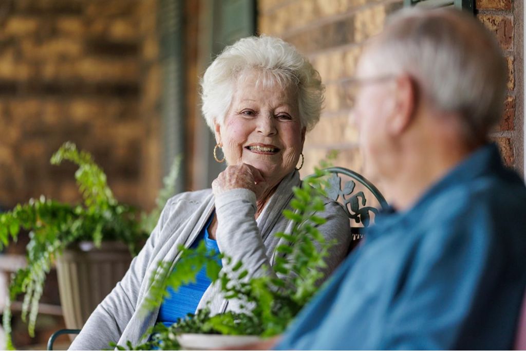 A smiling elderly woman sitting on a porch, looking happily at an older man seated next to her.