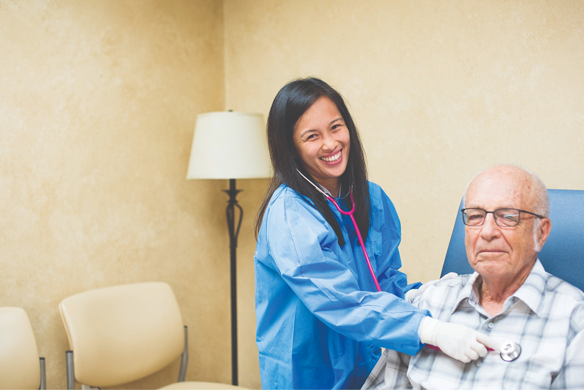 A smiling female healthcare worker in a blue gown and white gloves using a pink stethoscope to check the heartbeat of an elderly male patient seated in a clinic.