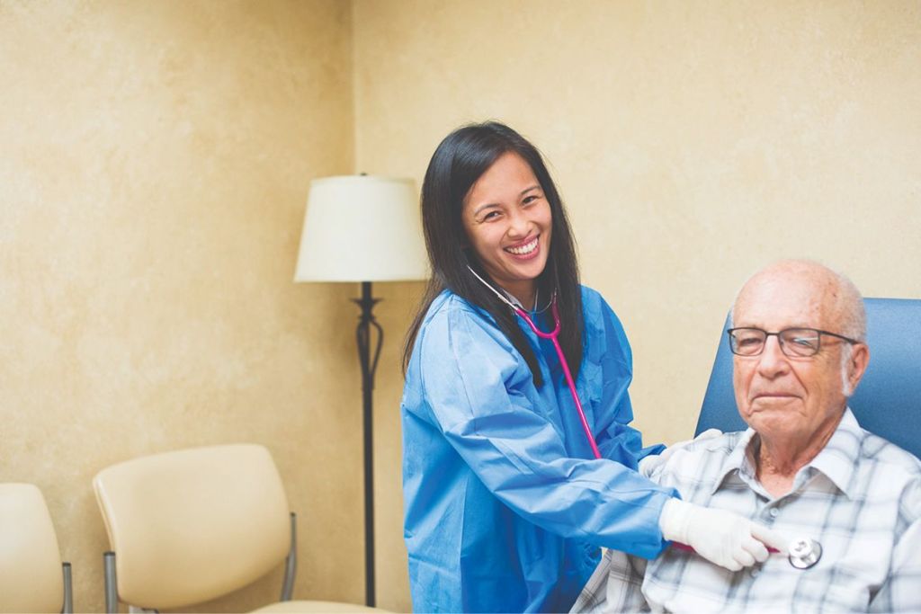 A smiling female healthcare worker in a blue gown and white gloves using a pink stethoscope to check the heartbeat of an elderly male patient seated in a clinic.
