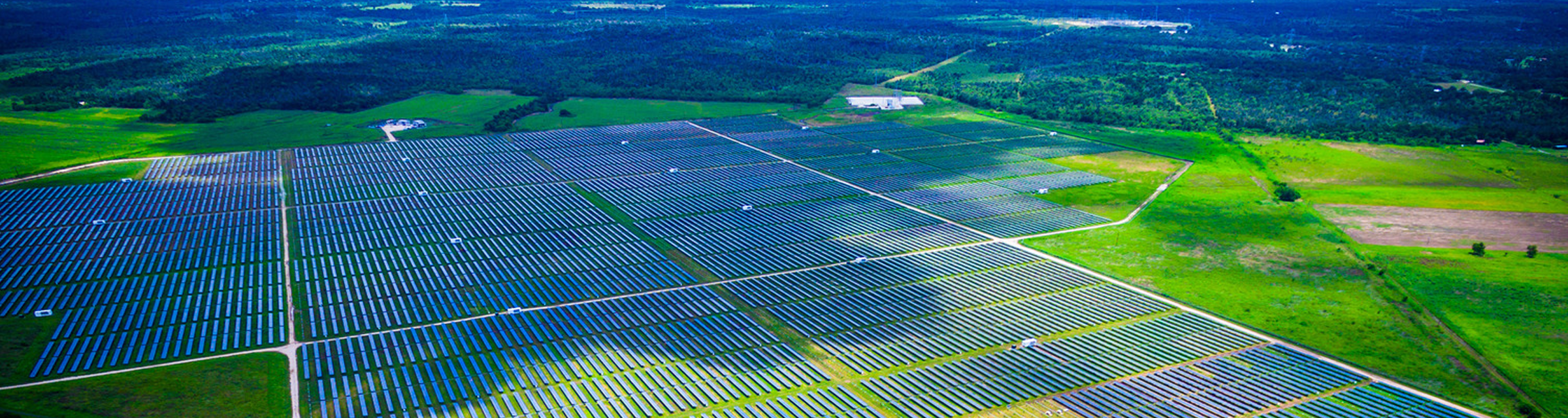 An aerial view of a large-scale solar panel farm situated in a dry, arid landscape with rolling hills and mountains in the background.